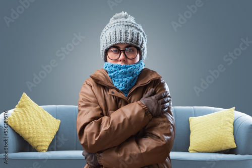 Young shocked woman in a knitted hat, coat and scarf is trying to keep warm, hugging herself. Gray background with sofa. The concept of energy crisis and winter season