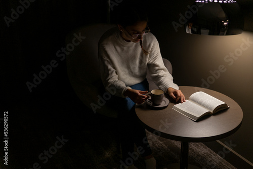 Woman reading a book in her room at night with a coffee. Concept of reading novels at night.