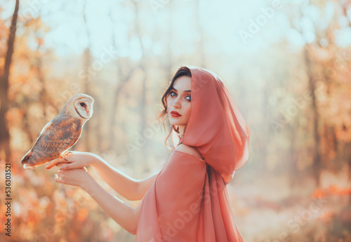 mystical fantasy woman walks in autumn orange forest, holds cute pet bird little barn owl on hand. Girl Princess. medieval old style clothig cape hood peach dress. bright make-up red lips pale skin, photo