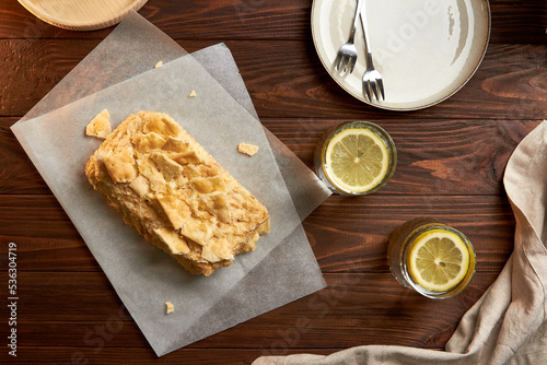 Piece of napoleon cake. Creamy vanilla slice dessert on a parchment paper. Layered baked pie with two glasses of water. Sweet food presentation. Copy space. Woody background. photo