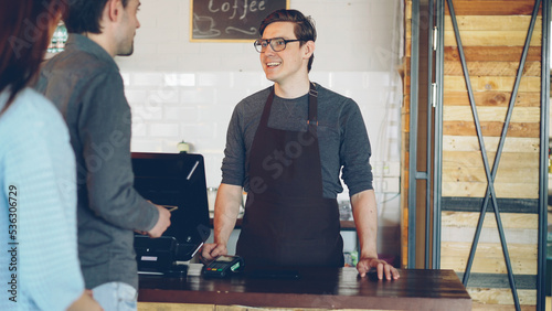 Cheerful friendly cashier is taking orders from customers standing in line, accepting payment and selling takeaway coffee.