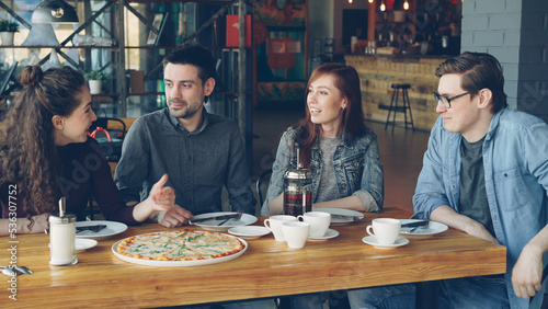 Cheerful young friends are talking and gesturing sharing news while sitting at table in modern cafe together. Big pizza, cups and plates, tables and chairs are visible.