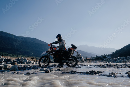 motorcyclist wearing a helmet rides an off-road motorcycle along ford of mountain river in beautiful landscape