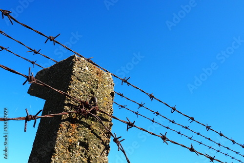 Barbed wire, double wire, metal tape with sharp spikes for barriers. Rusty barbed wire against the blue sky. The concept of war, restriction of rights and freedoms. Concrete pillar. photo