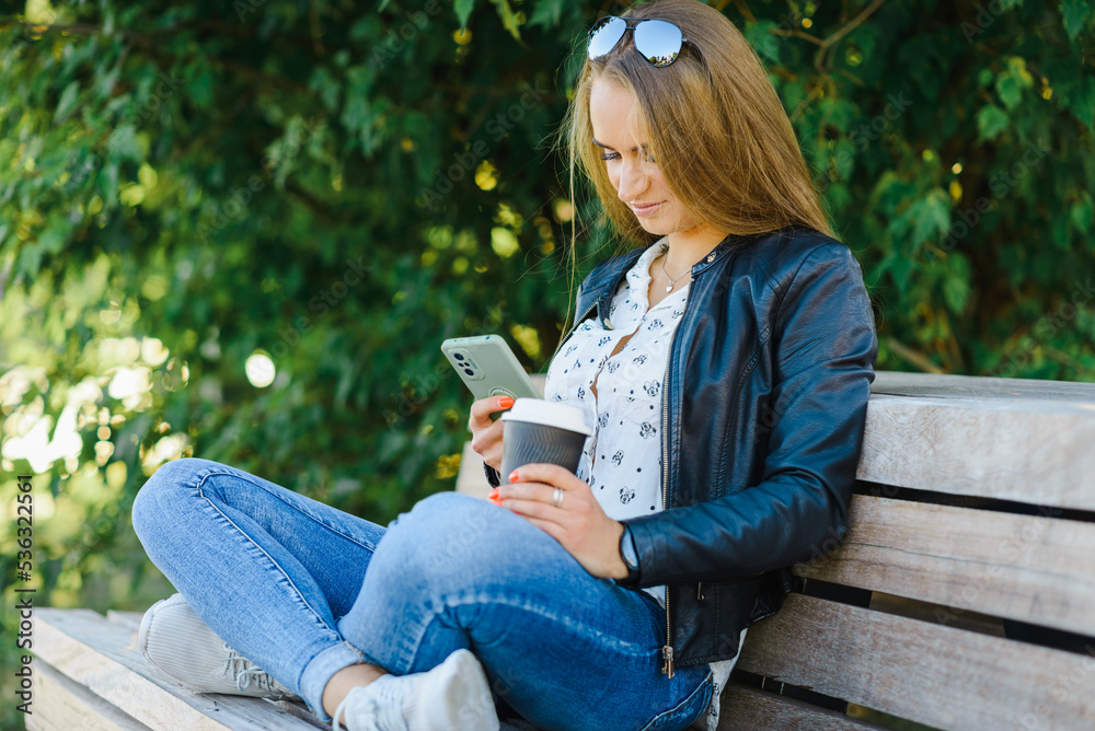 Obraz premium A smiling modern young woman sitting on bench in green park on summer day and talking on smartphone with coffee cup.Girl sitting with a phone and coffe at park.Klaipeda,Lithuania.09-02-2022.