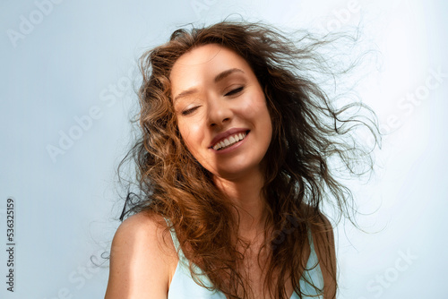 Portrait of happy young woman smiling in the sunshine on  blue background. Model with curly  hair and perfect skin. Haircare concept.