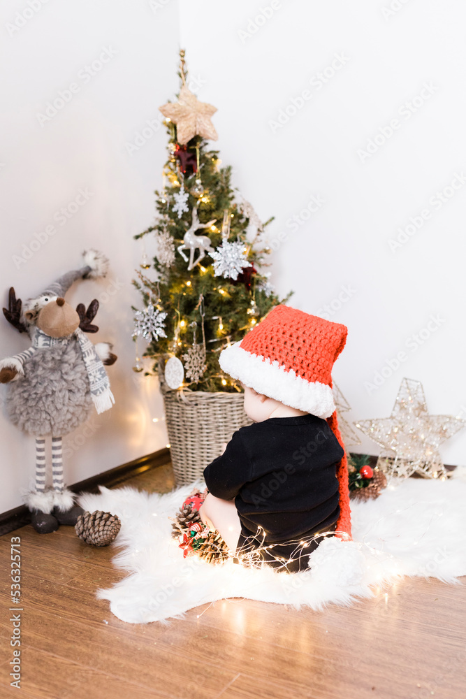 Obraz premium A baby boy is sitting in front of a Christmas tree, he is holding new year lights and wearing Santa Clause red hat