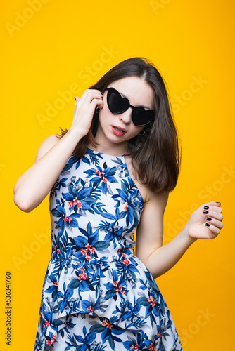Portrait of a young woman in a dress and sunglasses on a yellow background.