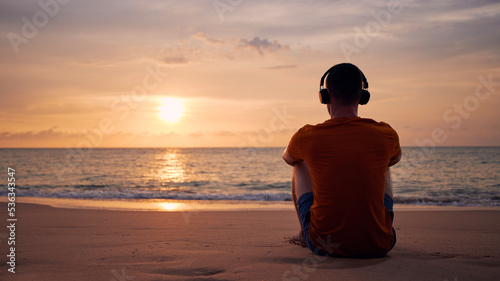 Rear view of man with headphones on beach. Serenity, contemplation and listening music at beautiful sunset..