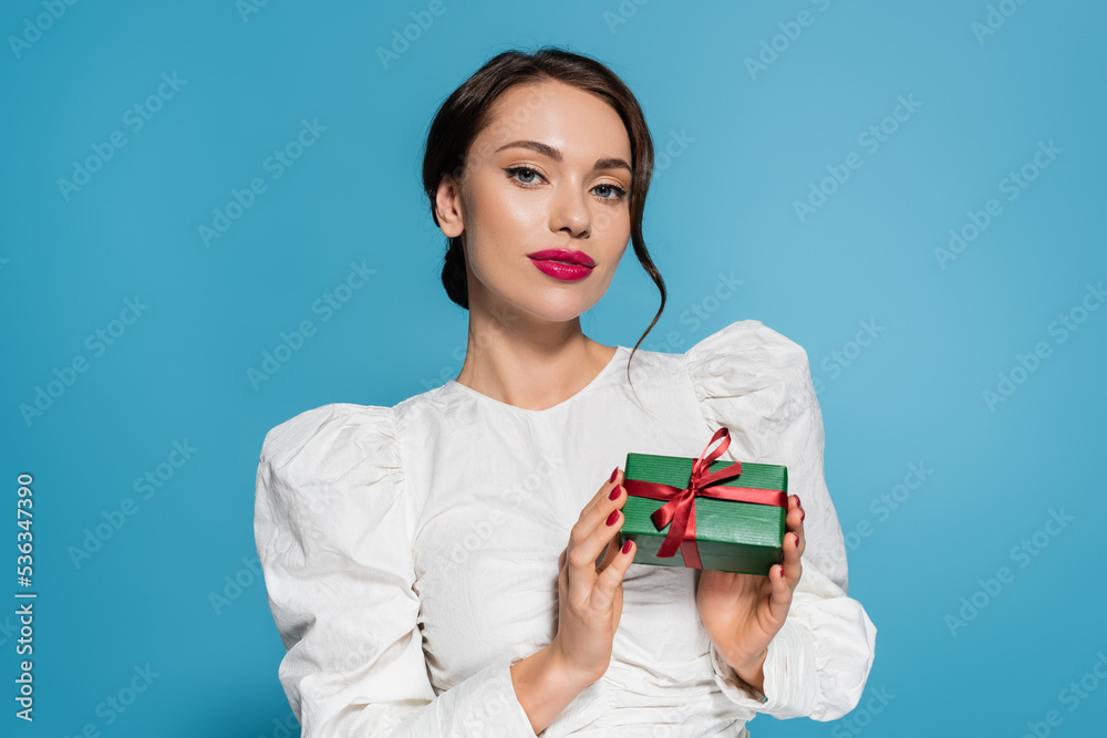 brunette young woman in white blouse holding wrapped present and pooing at camera isolated on blue. brunette young woman in white blouse holding wrapped present and pooing at camera isolated on blue.