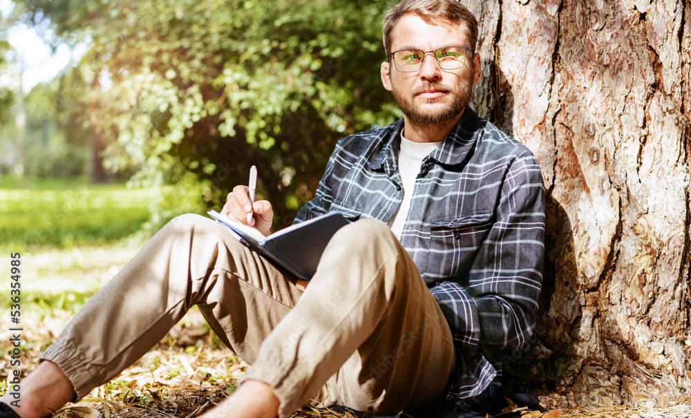 Fototapeta premium Man with eyeglasses wearing casual clothes writing in his diary while sitting in the park leaning on the tree.