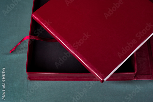 A large stylish box and an expensive premium burgundy photobook with a leather cover and white pages lie on a gray table indoors.