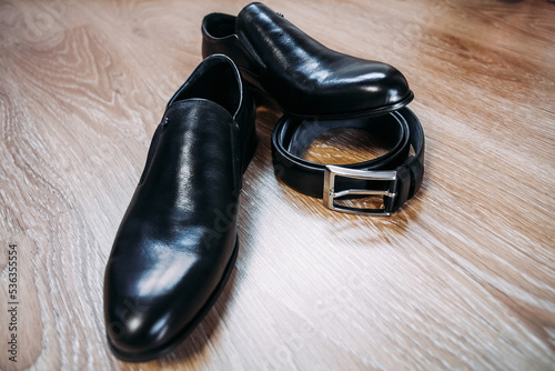 Black men's patent leather shoes for the groom and a leather black belt with an iron buckle lie on a wooden brown textured surface.