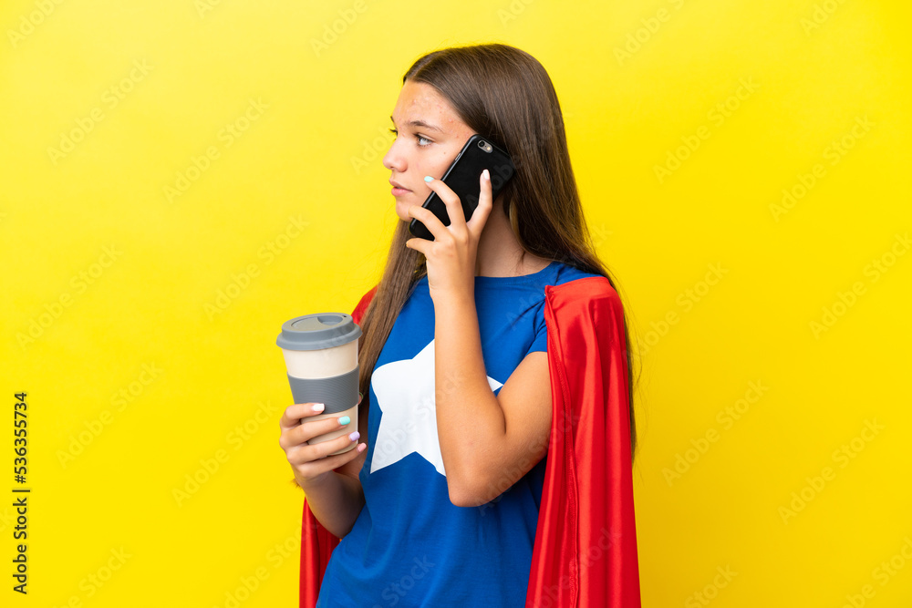 Little caucasian superhero girl isolated on yellow background holding coffee to take away and a mobile