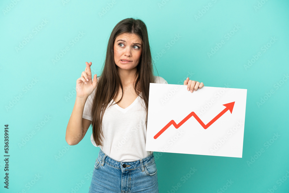 Young caucasian woman isolated on blue background holding a sign with a growing statistics arrow symbol with fingers crossing Young caucasian woman isolated on blue background holding a sign with a growing statistics arrow symbol with fingers crossing