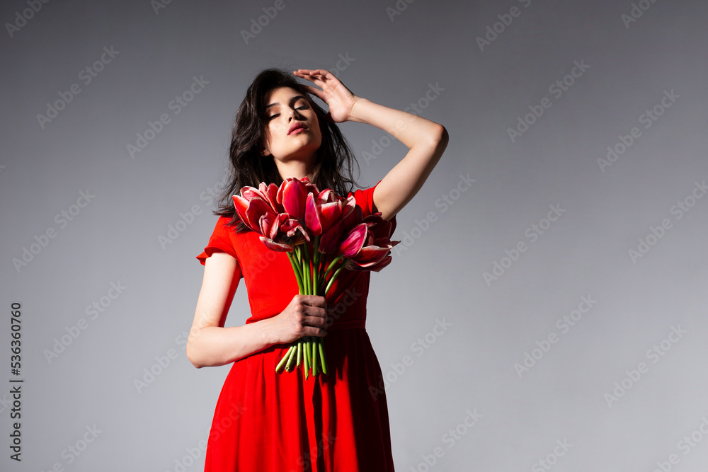 Attractive young woman in red dress holding a bouquet of tulips, posing sensual at camera, isolated grey background. Attractive young woman in red dress holding a bouquet of tulips, posing sensual at camera, isolated grey background.
