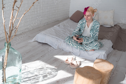 Young pink haired woman sitting in bed and reding ebook or social media or serfing internet using smartphone