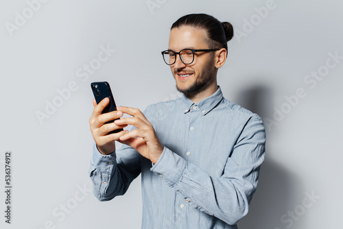 Studio portrait of young smiling man using smartphone on white background, wearing blue shirt and eyeglasses.