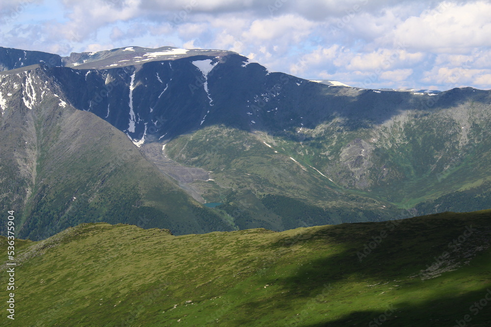 Fototapeta premium The slopes of huge mountains with grass and rocks in summer in sunny weather, on the far slope you can see a lake, in some places there is snow, a sky with clouds