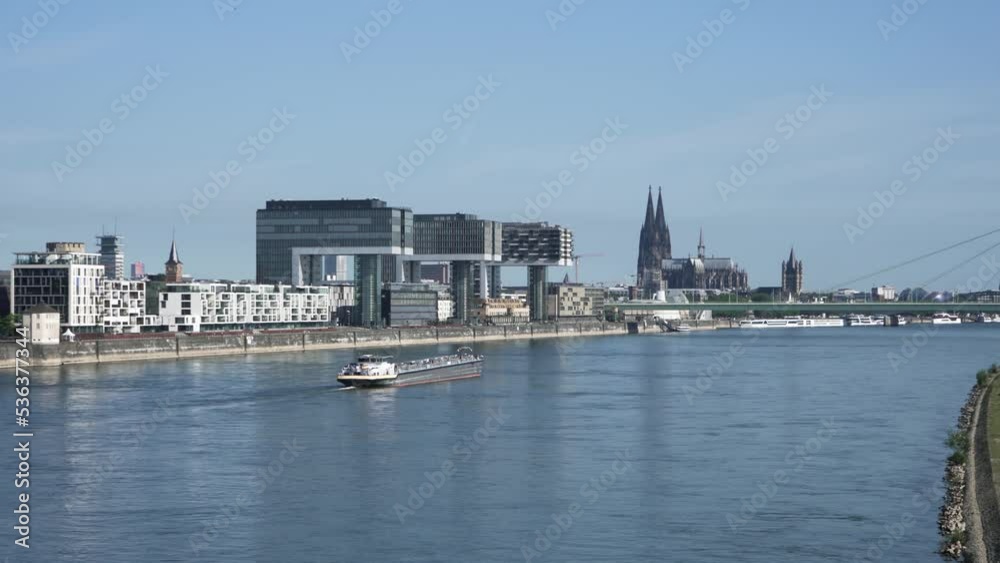 Boat at Rhine river with modern office buildings and Dom cathedral standing on riverbanks in Cologne.
