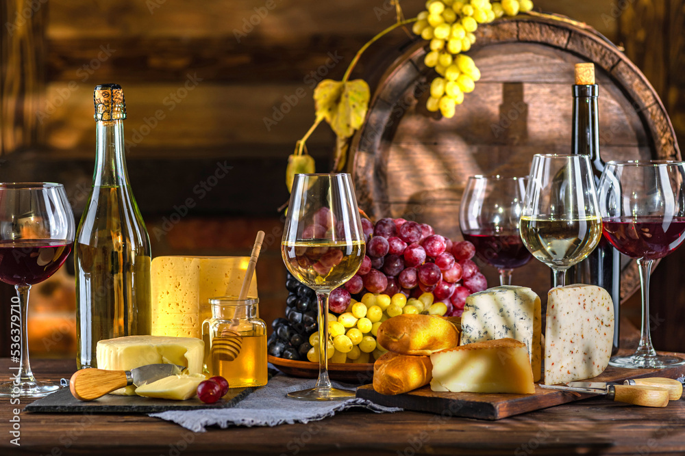 Cheese and wine tasting still life. Glass of wine, bottle of wine, grape in front of the rustic wooden barrels for beverage in a dark wine cellar. Nuts, honey, dipper. Empty copy space, mock up