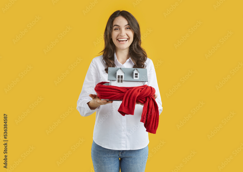 Joyful woman holds toy house wrapped in warm scarf as symbol for heating system or cold snowy winter. Isolated on orange background. Building insulation, heating season, warm up and protect concept. Joyful woman holds toy house wrapped in warm scarf as symbol for heating system or cold snowy winter. Isolated on orange background. Building insulation, heating season, warm up and protect concept.