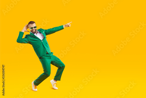 Full body happy funny smiling joyful handsome young man in trendy stylish green suit and sunglasses having fun, dancing isolated on yellow color background and pointing finger away at blank copy space