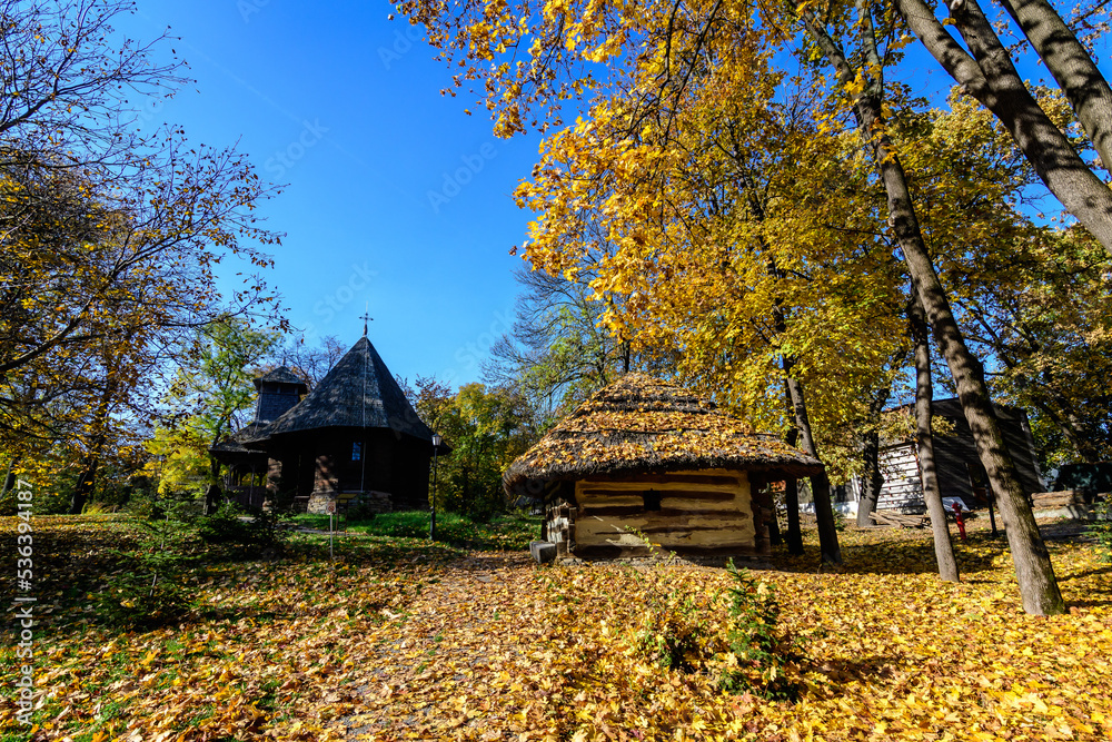 Traditional Romanian house surounded with many old trees with green, yellow, orange and brown leaves in Village Museum in Herastrau Park in Bucharest, Romania in a sunny autumn day.