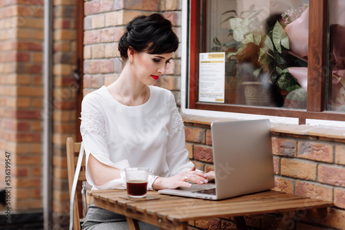 Young businesswoman working on laptop at cafe on terrace in urban city, stylish outfit with white blouse and skirt. Coffee break, business concept. Working outdoors. Remote job. © Andriy Medvediuk