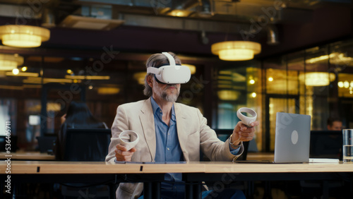 Male office worker wearing VR headset and using wireless controllers and laptop, gesturing, watching data in virtual reality. Work in modern office of hi-tech company. Cyberspace digital technology