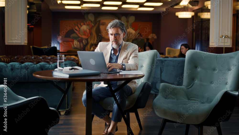 Mature businessman using laptop, working online on startup project, business strategy, analyzing papers and documents in office cafe. Worker of E-commerce marketing company Mature businessman using laptop, working online on startup project, business strategy, analyzing papers and documents in office cafe. Worker of E-commerce marketing company