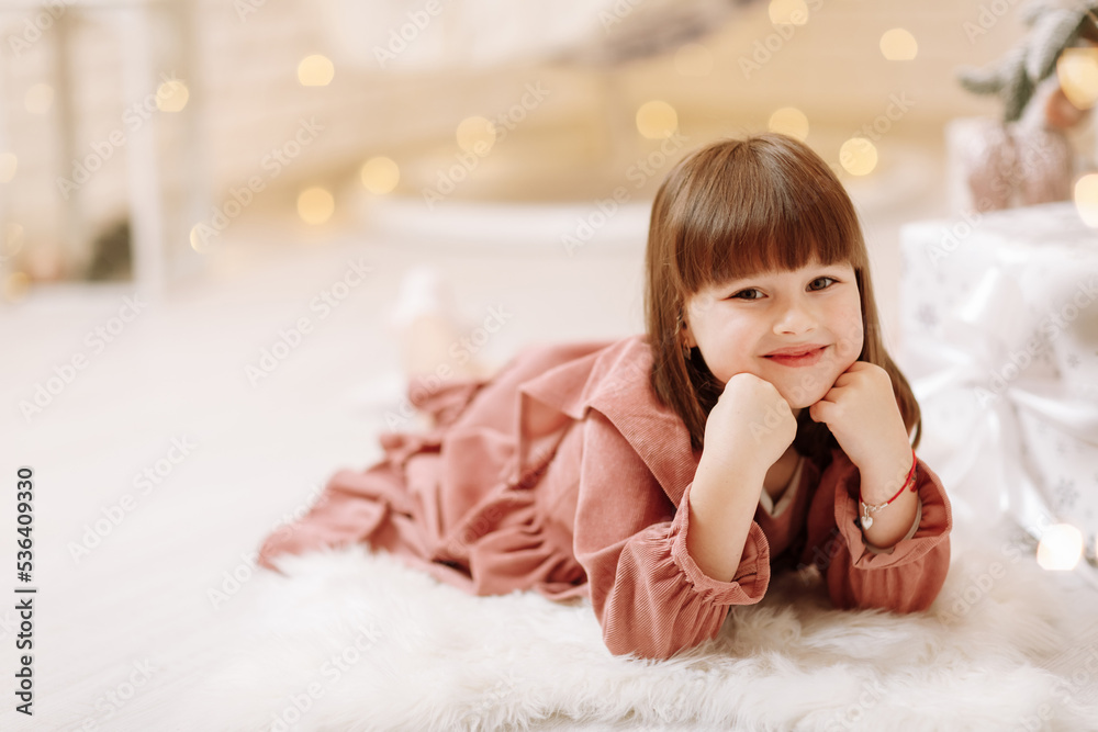 Fototapeta premium little cute child girl in pink dress lying on white furry carpet in decorated Christmas room interior and looking in camera. Beautiful bokeh. Happy family New Year, Xmas concept