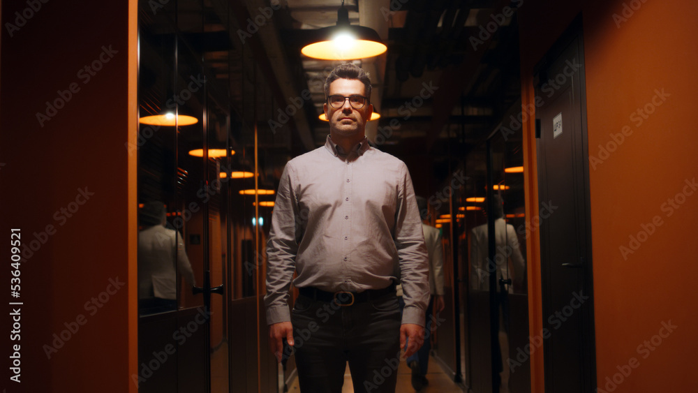 Happy male office worker in suit and glasses standing in office corridor, smiling, looking at camera. Successful mature businessman in hallway of marketing company modern business center