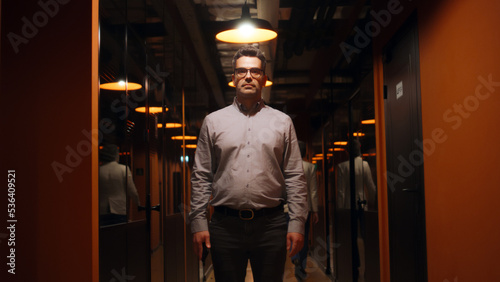 Happy male office worker in suit and glasses standing in office corridor  smiling  looking at camera. Successful mature businessman in hallway of marketing company modern business center