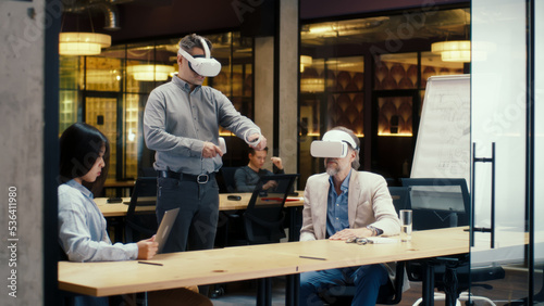 Multi-ethnic office workers testing VR headsets together, using tablet computer and wireless controllers, watching data, working in modern office of hi-tech company. Cyberspace digital technology