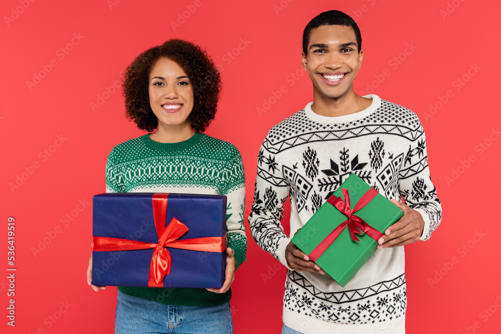 joyful african american couple with green and blue gift boxes looking at camera isolated on red joyful african american couple with green and blue gift boxes looking at camera isolated on red