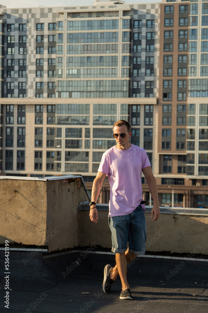 A handsome guy in denim shorts and a pink t-shirt poses beautifully in the city