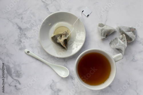 Cup of tea, tea bag on saucer and spoon on table. Green herbal tea in mug on white marble background. Top view. Traditional teatime. Teabag in cup 