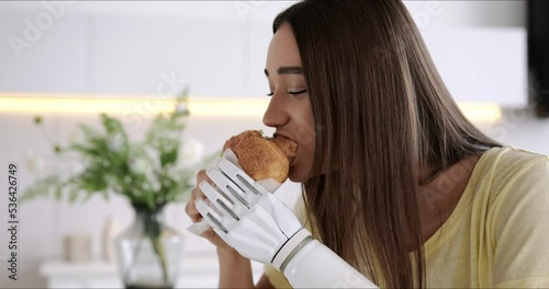 Girl using prosthetic arm while eating at home, woman with disability holds croissant in artificial prosthetic limb bionic hand photo