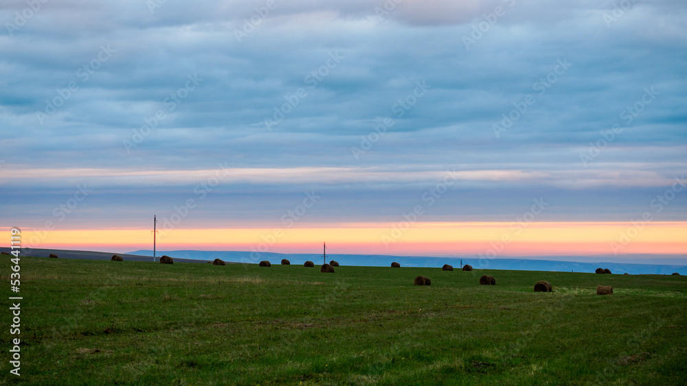 Fototapeta premium Sunset colored sky over green fields with with hay bales and utility poles