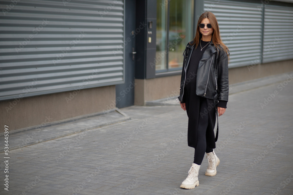 Stylish beautiful pregnant girl walks along a city street.Wearing a black leather jacket, a black long dress and white high boots and black sunglasses. Stylish beautiful pregnant girl walks along a city street.Wearing a black leather jacket, a black long dress and white high boots and black sunglasses.