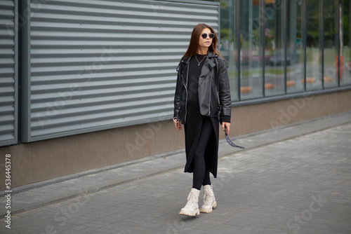 Stylish beautiful pregnant girl walks along a city street.Wearing a black leather jacket, a black long dress and white high boots and black sunglasses.