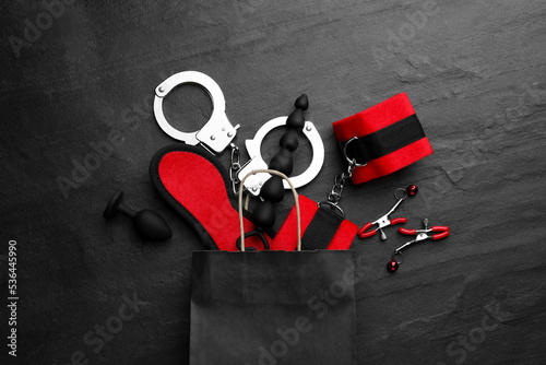 Paper shopping bag with different sex toys on black table, flat lay