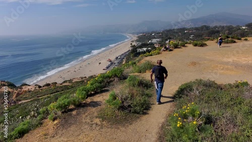 aerial footage of an African American man wearing a backpack walking around on the tops of the cliffs at the beach surrounded by red dirt and lush green grass and plants with blue ocean water