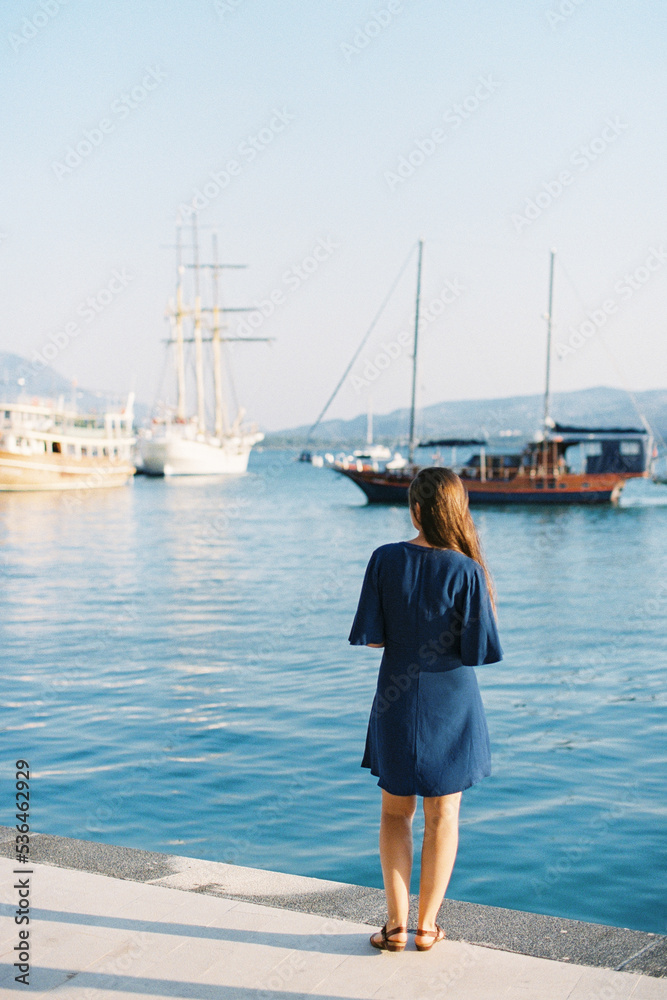 Woman in a blue dress stands on the pier and looks at the ships