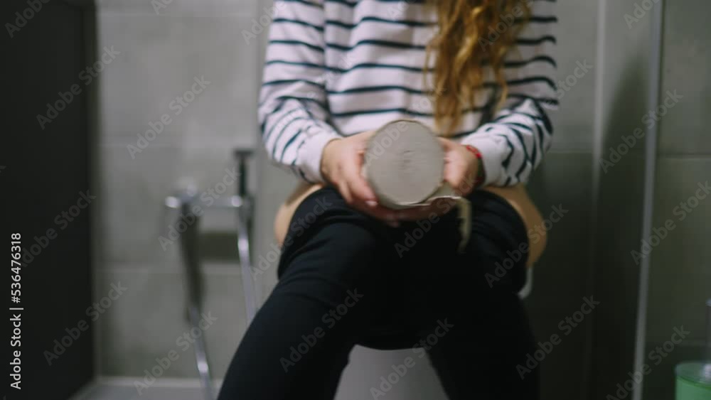 Low section of woman body sitting on toilet and suffers from diarrhea and stomachache. Female can't poop because of hemorrhoids piles. Strained girl holding and shaking toilet paper in hands.