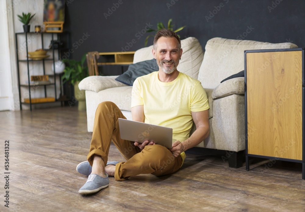 Portrait of handsome smiling man 45s wearing smart casual dress sitting on floor with laptop computer at living room with black wall background. Man lean on sofa