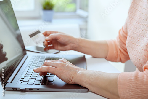 Mature woman works at home on a laptop. She is holding credit card in her hand. Closeup of hands  casual wear. Online shopping  work from home and freelance concept.