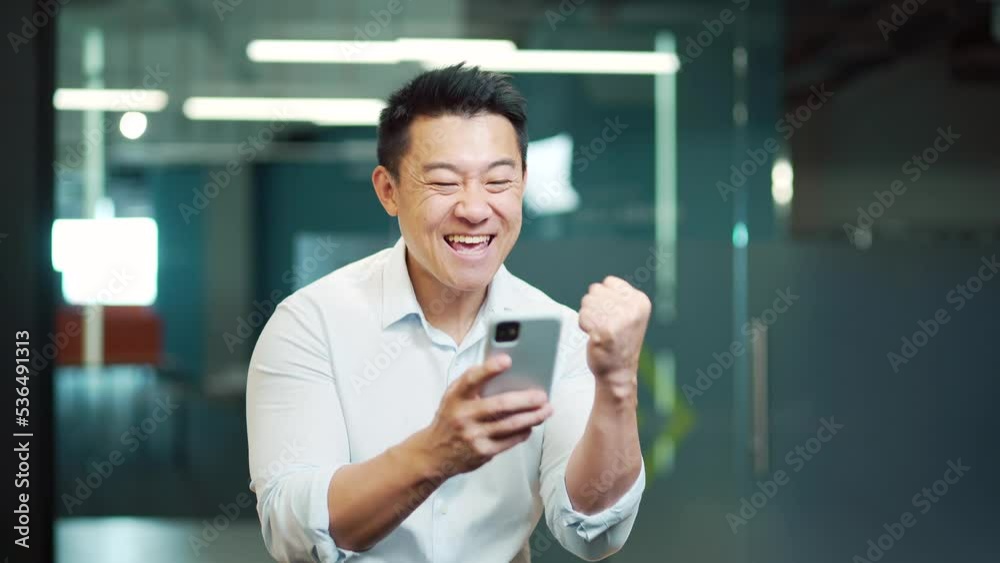 Happy asian man employee enjoys success read good news in smartphone. businessman win mobile online bet bid money financial prize victory receive offer opportunity on smart phone in the office indoor Happy asian man employee enjoys success read good news in smartphone. businessman win mobile online bet bid money financial prize victory receive offer opportunity on smart phone in the office indoor