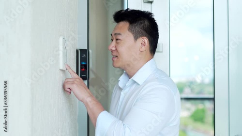 Asian man, calling rings the intercom of the house, pressing the button, waiting for the door to open. Male pushing the button and talking doorbell with camera near entrence electronic code lock photo